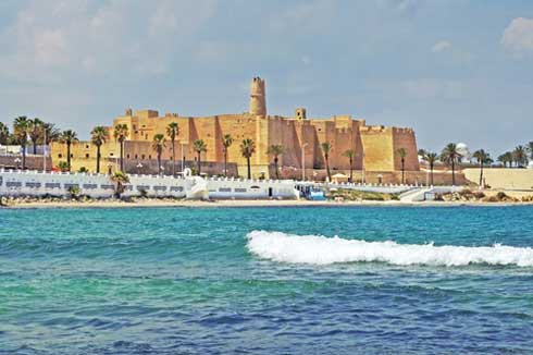 Ocean waves crash on a beach, blue skies, palm trees, sandstone castle. Summer car maintenance tips.