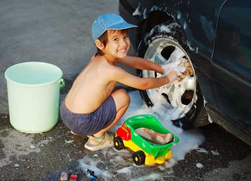 Boy washing tires in summer with toy truck sponge and soapy suds.