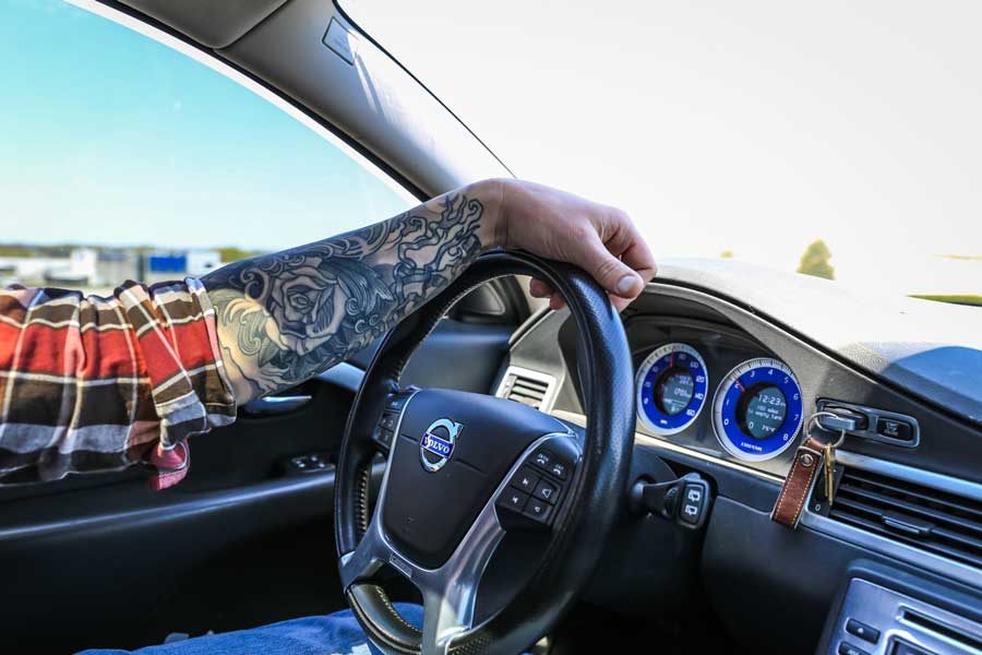 Man's tattooed arm on steering wheel practicing good ergonomics.